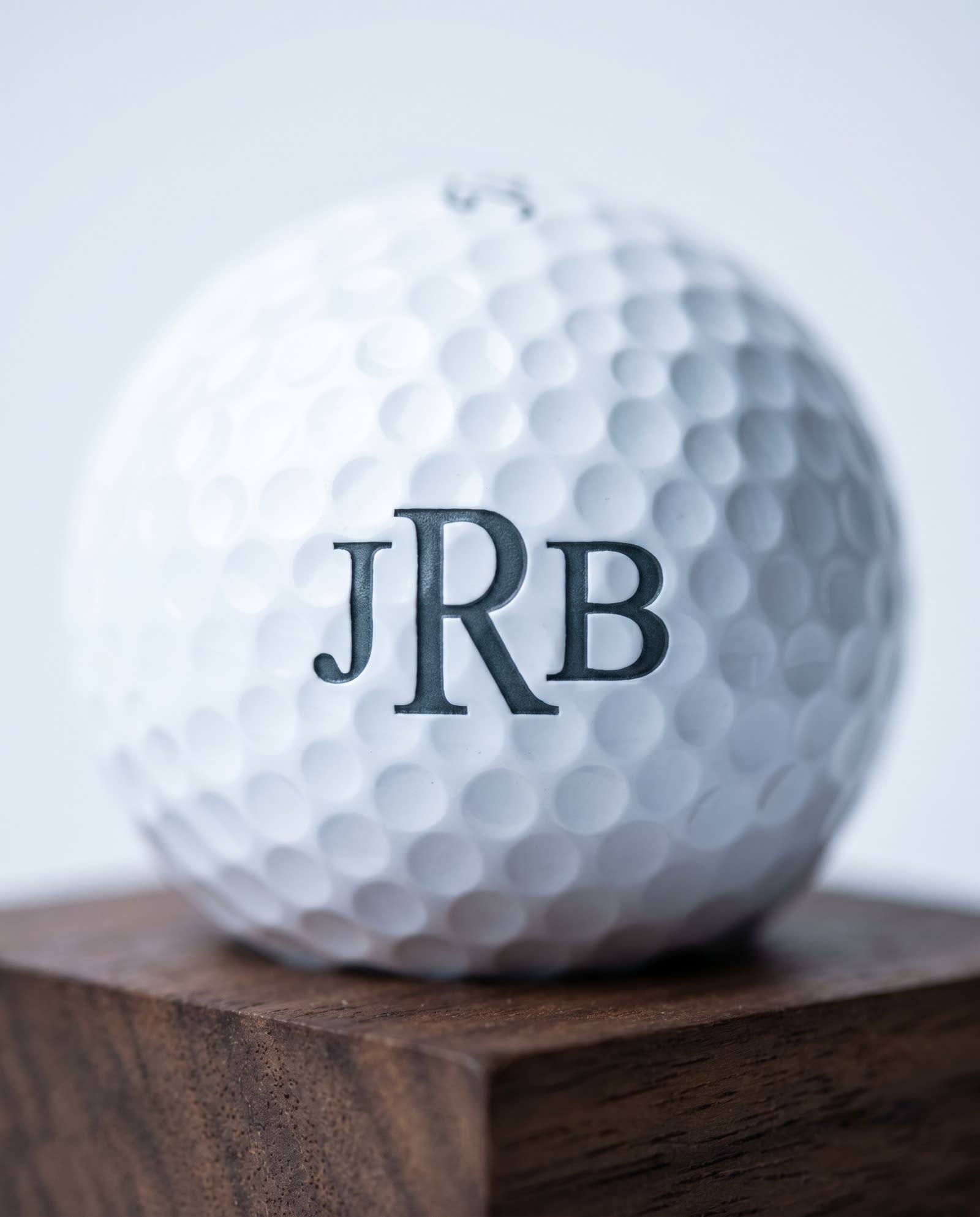Extreme macro of a single premium white golf ball UV-laser-marked with a deep three-letter serif monogram JRB on the urethane cover, photographed on dark walnut with shallow depth of field