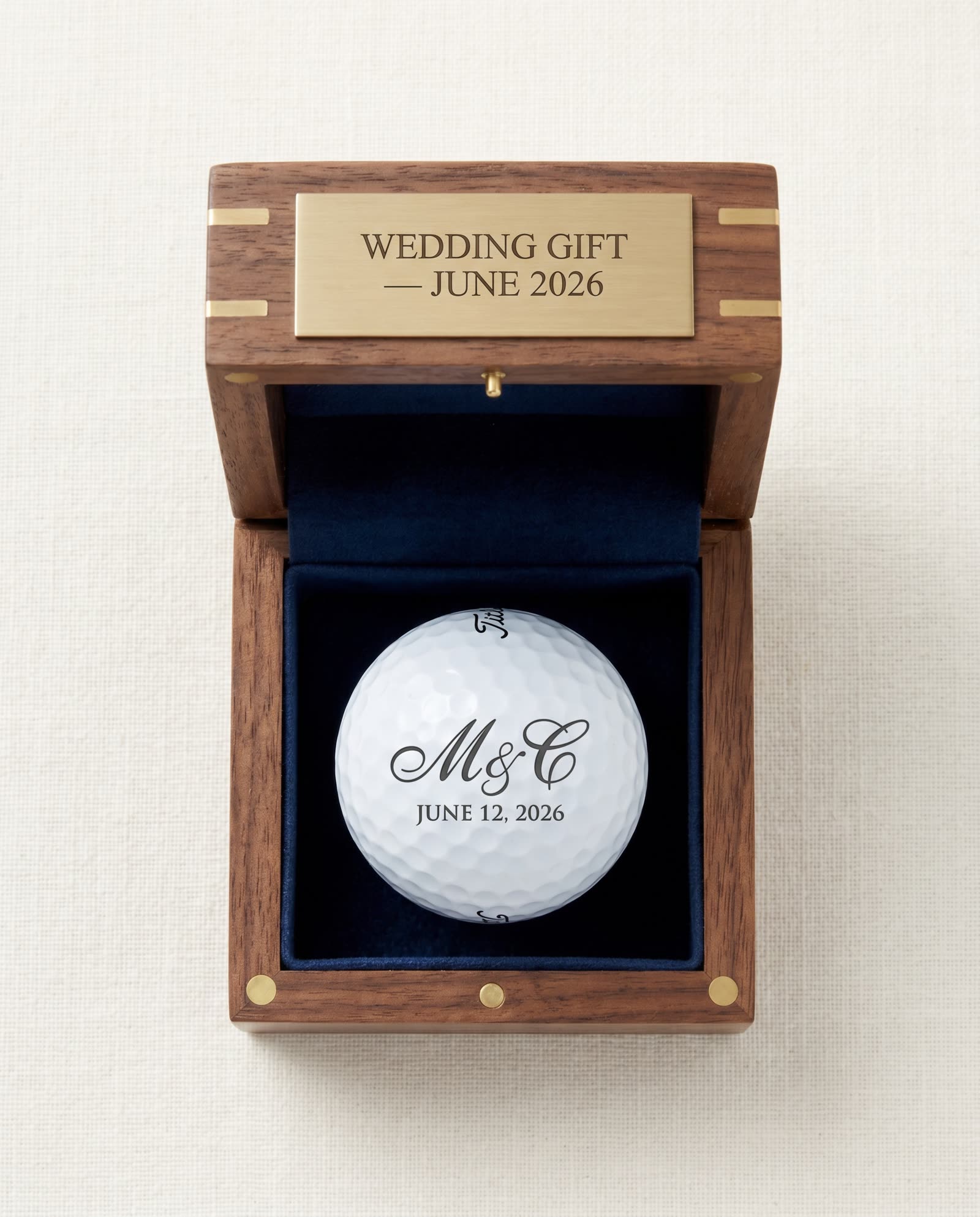 Custom golf ball wedding gift presentation — single UV-laser-marked Titleist ball with couple's monogram M & C and date JUNE 12 2026 displayed in a navy velvet-lined walnut box with brushed brass nameplate engraved WEDDING GIFT JUNE 2026