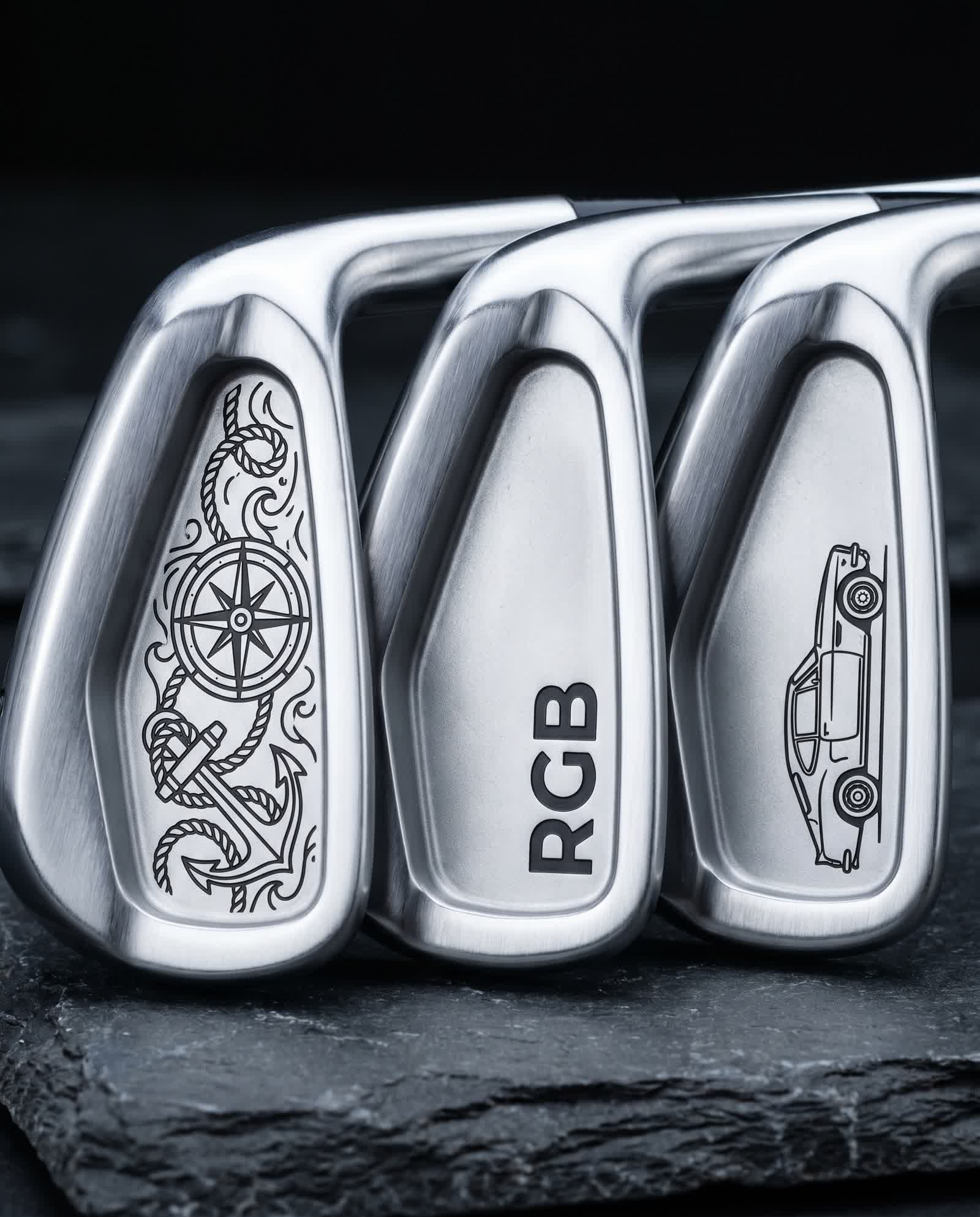 Three premium forged golf irons side by side, cavity backs laser black-annealed — left with a nautical anchor and compass-rose motif, center with bold RGB lettering, right with a classic car side-profile — flat dark marks on brushed satin steel, on dark slate in cool studio light