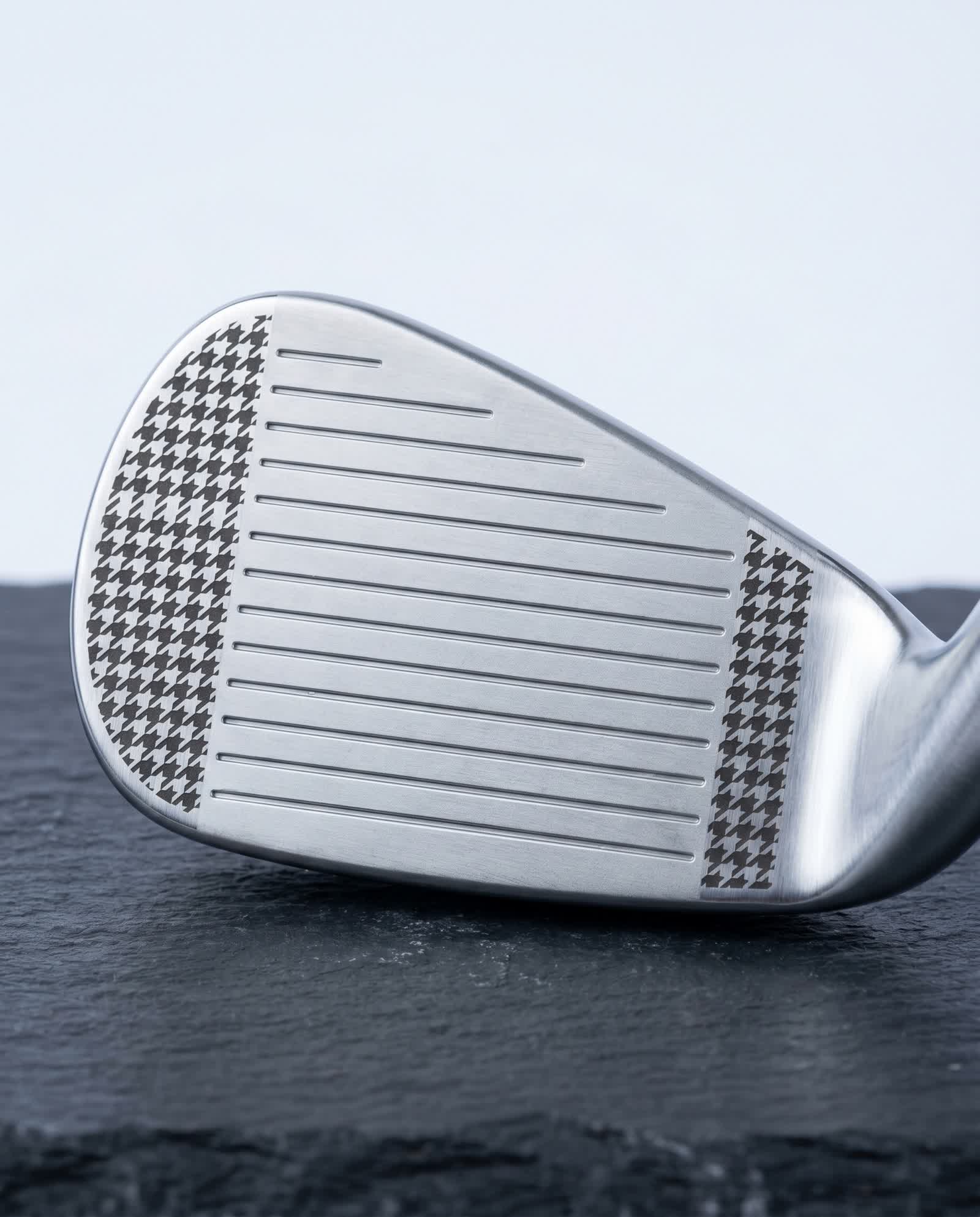 Macro of a premium golf iron face black-annealed with a crisp houndstooth pattern flanking the scoring grooves, flat dark marks on brushed satin steel, on dark slate in cool studio light