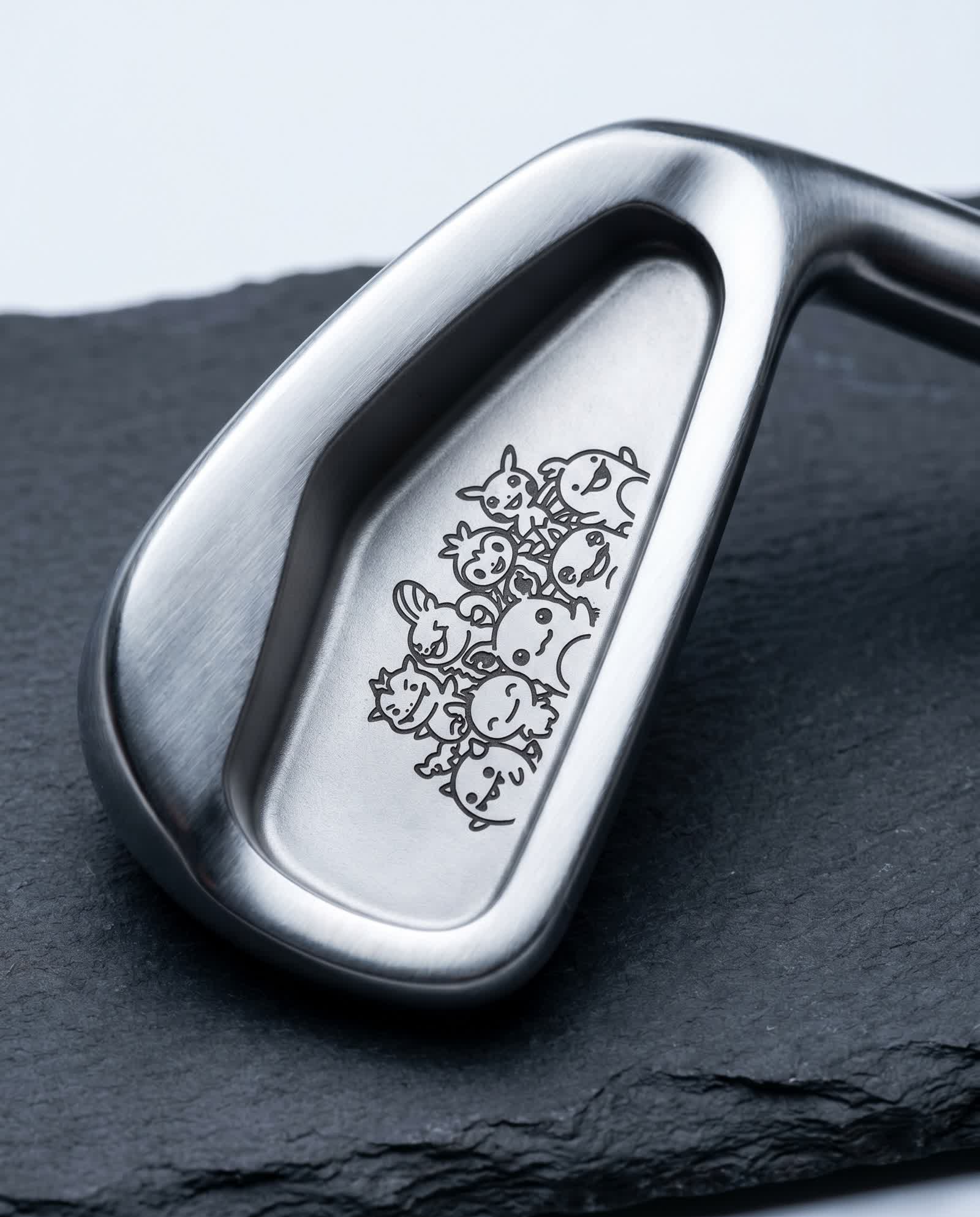 Macro of a premium golf iron black-annealed with a cluster of cartoon creature characters in line-art, flat dark marks on brushed satin steel, on dark slate in cool studio light