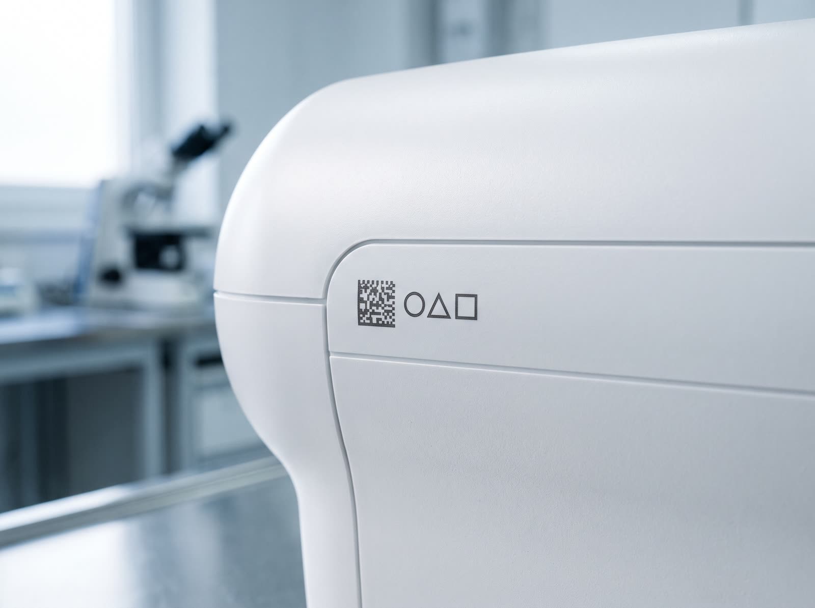 White medical-grade plastic housing of a laboratory device, UV-laser-marked directly in the plastic with a crisp dark data-matrix code and a row of simple pictogram symbols, no label or ink, with a bright clinical lab soft-focused behind