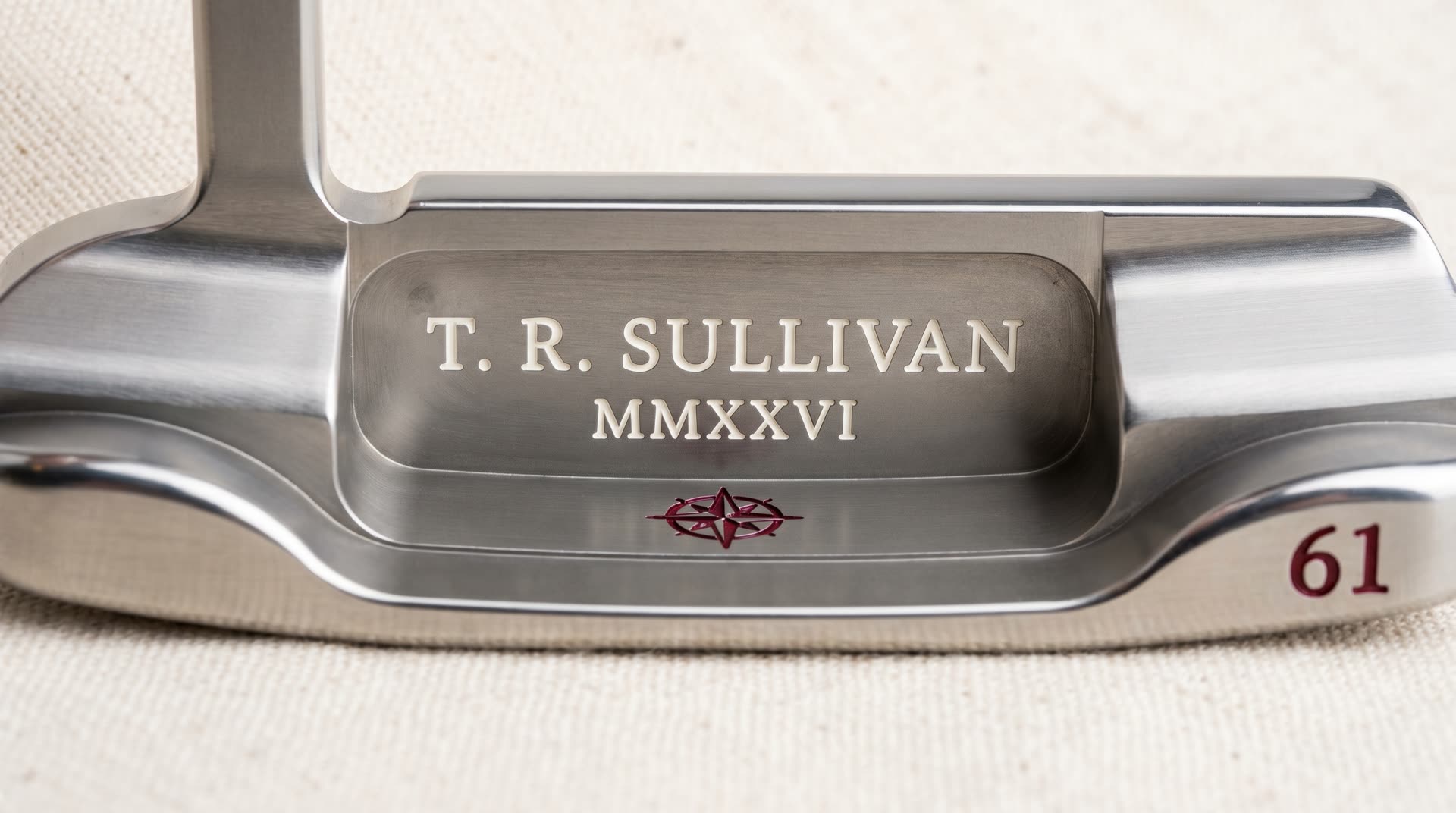 Macro of a premium milled blade putter cavity with custom multi-line engraving — recipient name T. R. SULLIVAN in serif, tournament year MMXXVI, decorative compass rose, and winning score 61 — two-color paint fill in bone white and deep crimson, shot on cream linen in cool studio light
