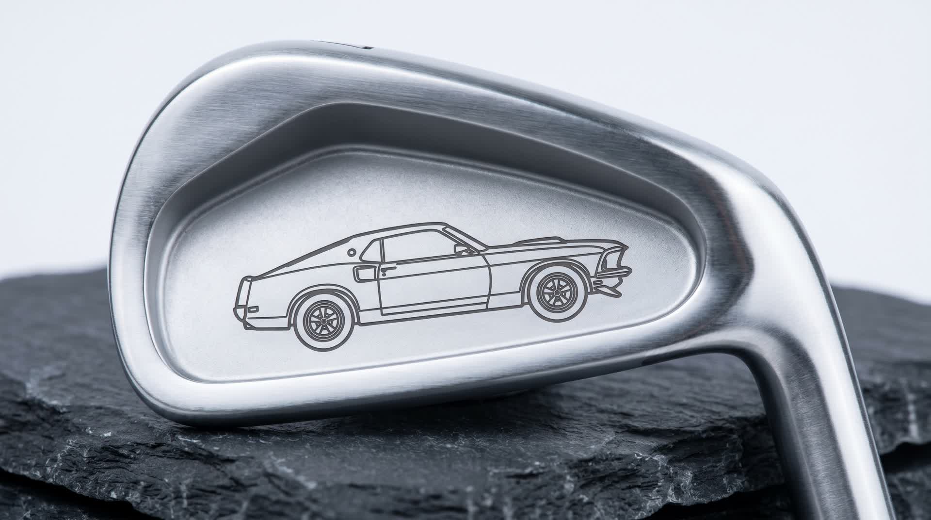 Macro of a premium golf iron cavity laser black-annealed with a side-profile illustration of a classic 1969 American muscle car in fine line-art, flat dark marks on brushed satin steel, shot on dark slate in cool studio light