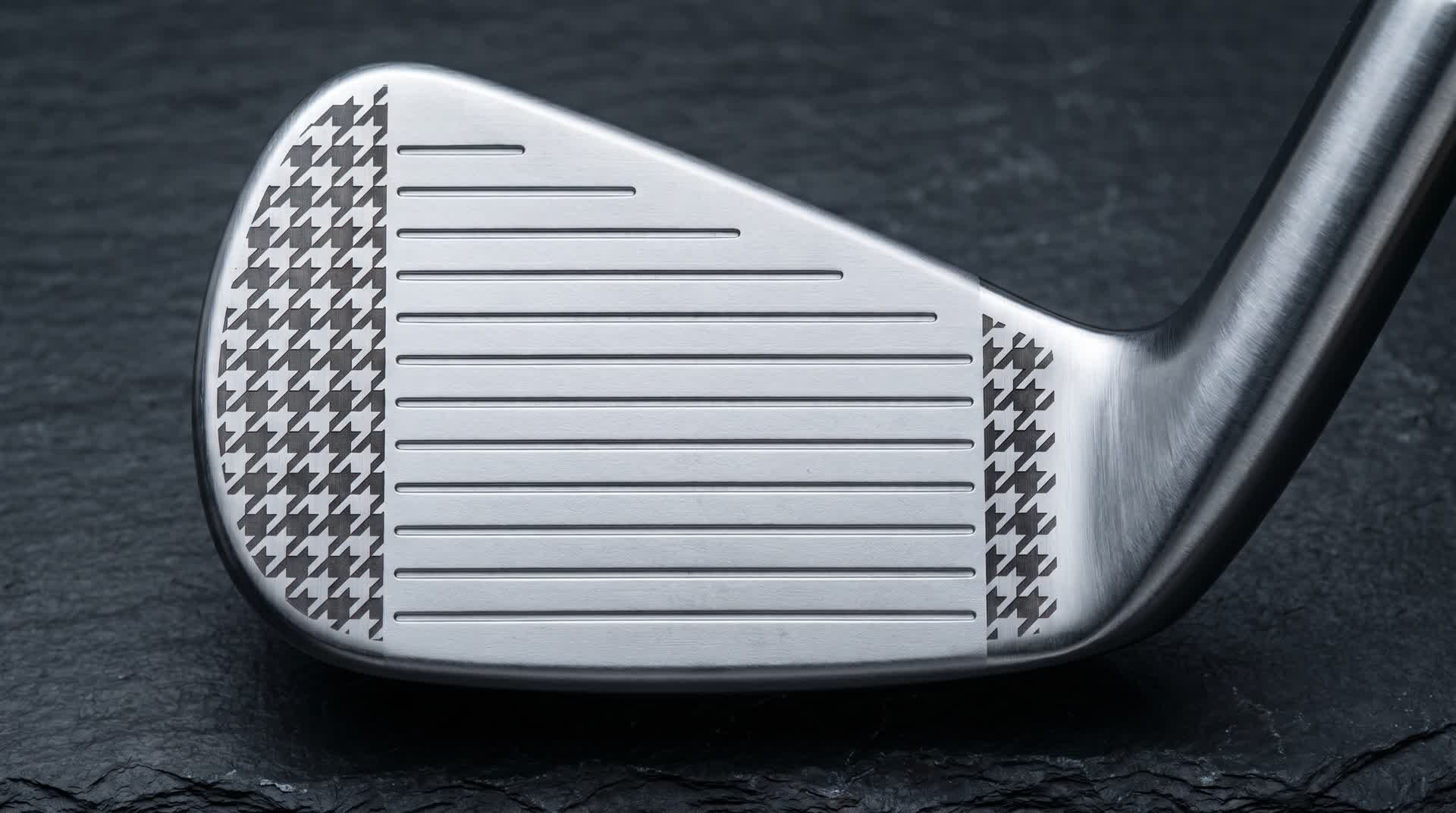 Macro of a premium golf iron face shown straight-on — horizontal scoring grooves across the center, with a crisp houndstooth pattern laser black-annealed on the heel-side and toe-side flats flanking the grooves, on dark slate in cool studio light